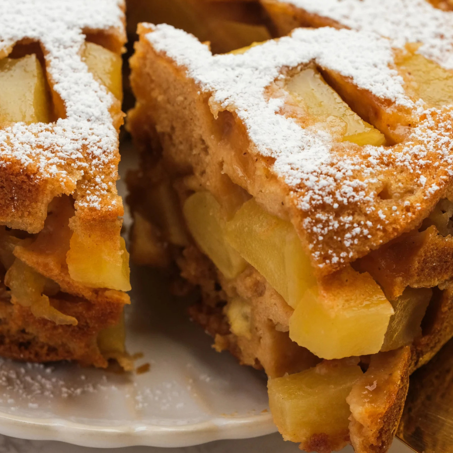 Close-up of a sliced apple cake or fruit cake on a white plate, dusted with powdered sugar and showing pale yellow apple pieces inside.