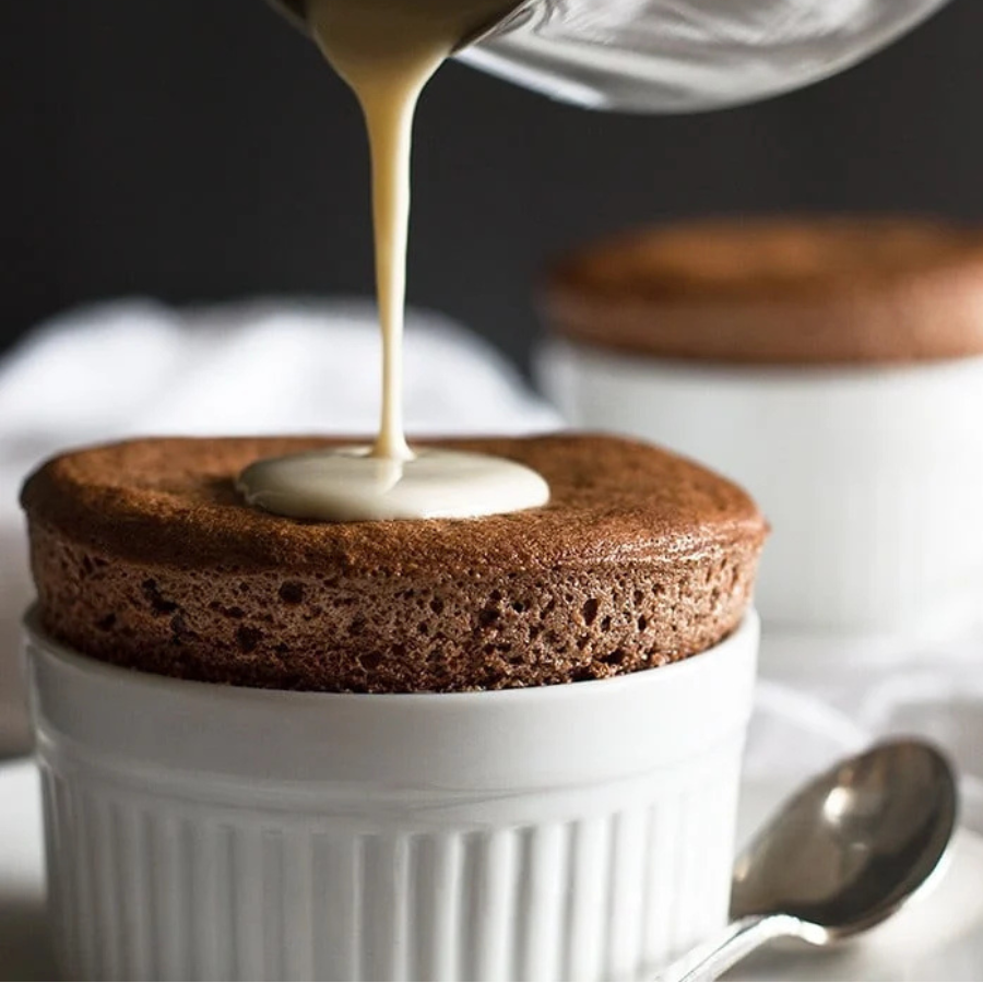 Chocolate souffle being drizzled with cream in a white ramekin.