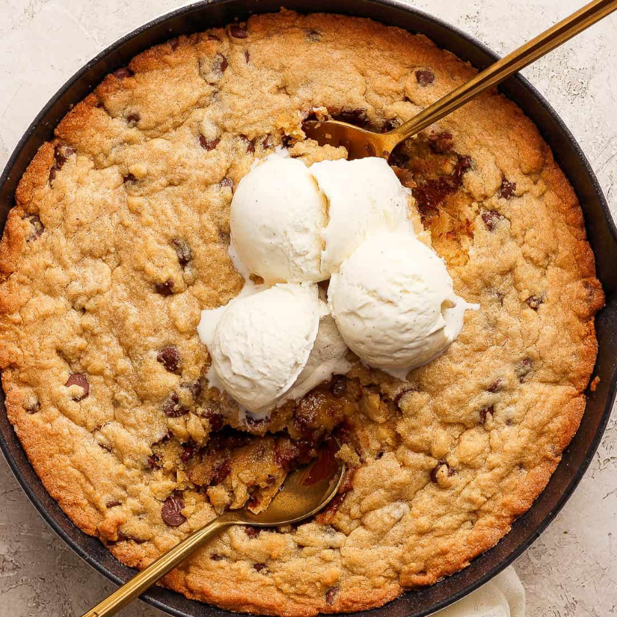 A chocolate chip skillet cake topped with three scoops of vanilla ice cream and gold spoons, photographed from above on a light stone surface.
