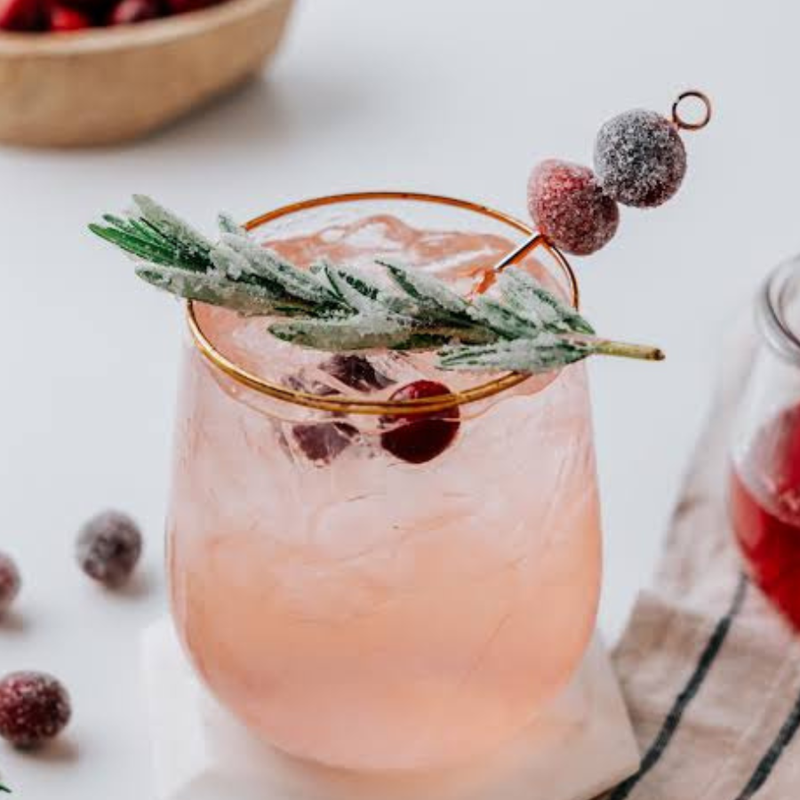 A pink cocktail in a clear stemless glass with a gold rim, garnished with a frosted rosemary sprig and red cranberries on a pick, set on a light tabletop with a napkin and additional cranberries nearby.