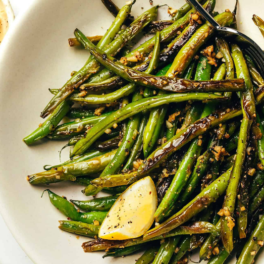 Close-up of cooked green beans with charred edges, garlic, and a lemon wedge on a white plate.