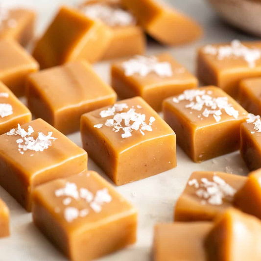Close-up of several square caramel candies topped with coarse white sea salt, arranged on a light surface.