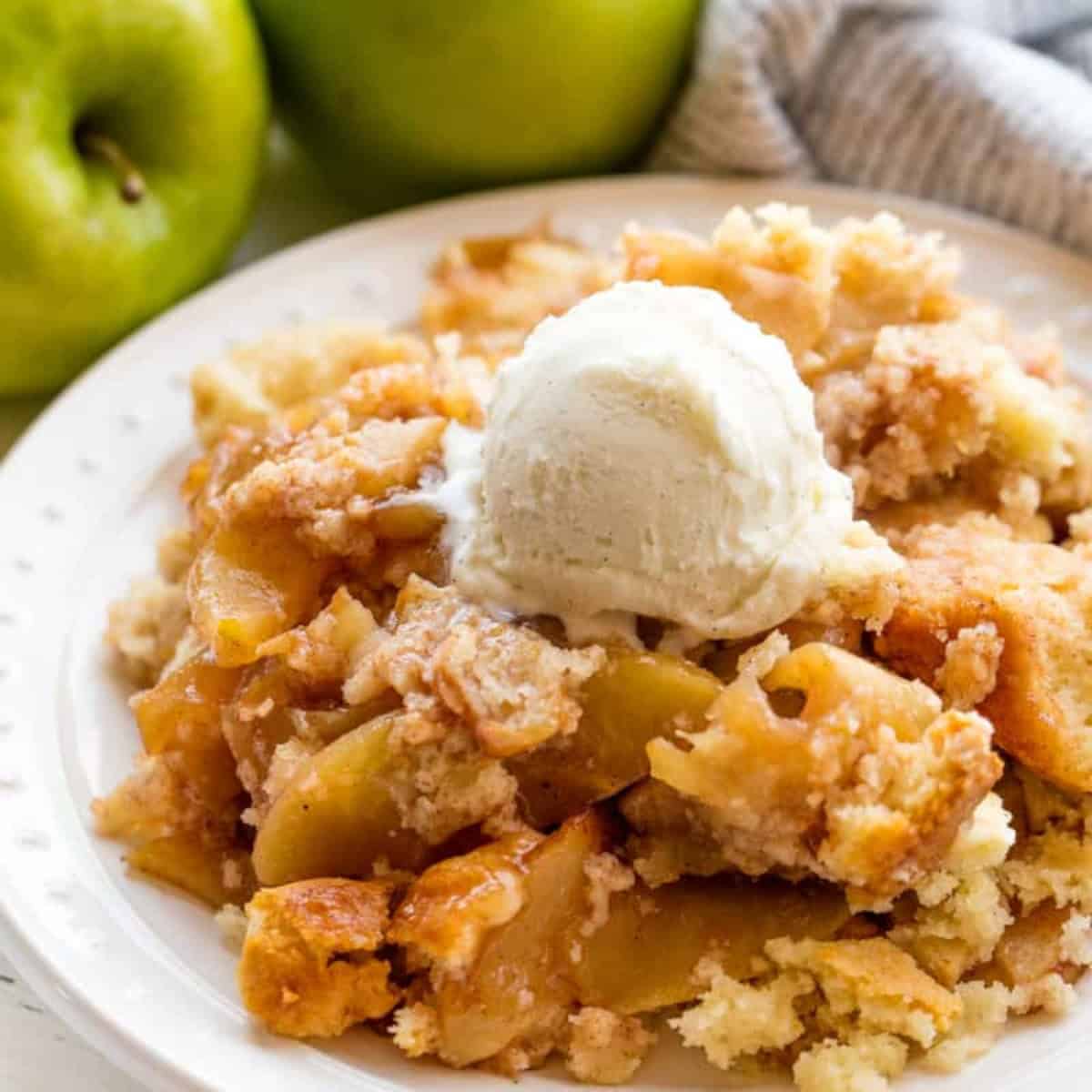 A close-up of apple crisp on a white plate, topped with a scoop of vanilla ice cream, with whole green apples and a striped cloth in the background.
