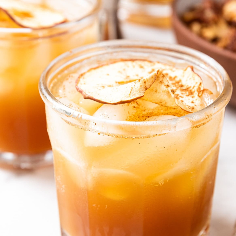 A close-up of an iced apple cider drink in a clear glass, topped with dried apple slices and a dusting of cinnamon, with a second glass blurred in the background.