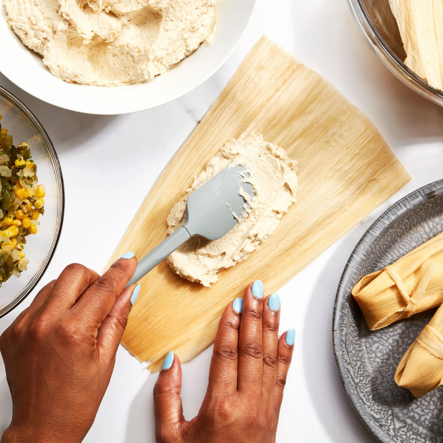 Top-down view of hands preparing a corn tamaño, spreading beige filling onto a corn husk with a gray spatula, surrounded by bowls of ingredients and finished tamaños on a white surface.