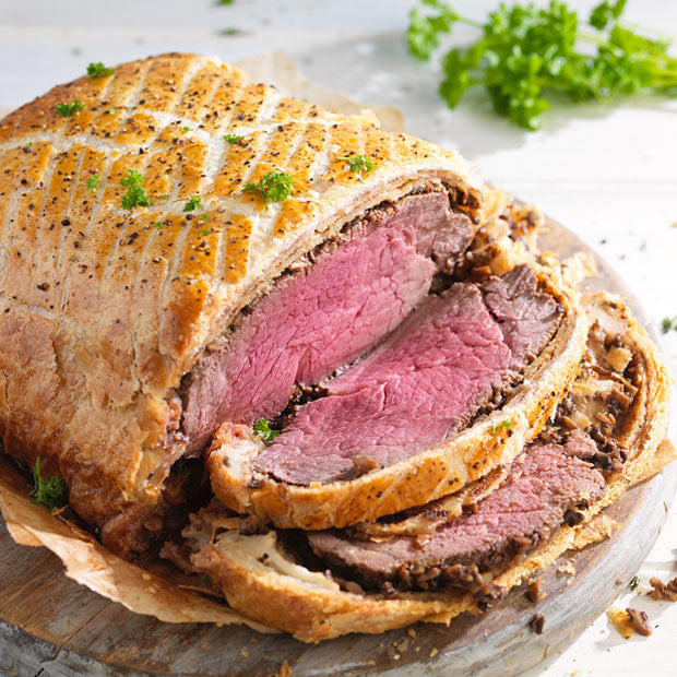 Sliced beef wellington on a wooden board with herbs in the background