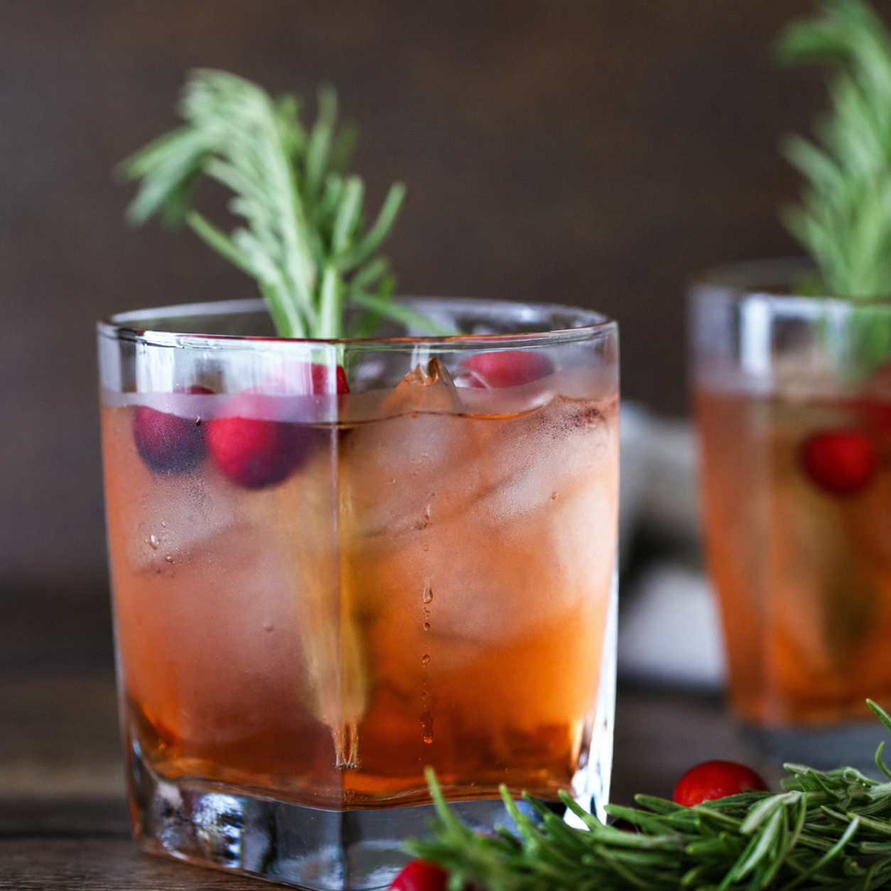 A lowball glass filled with an amber-orange cocktail, large ice cubes, and garnished with a green rosemary sprig and red cranberries, set on a dark wooden surface with a second blurred drink in the background.