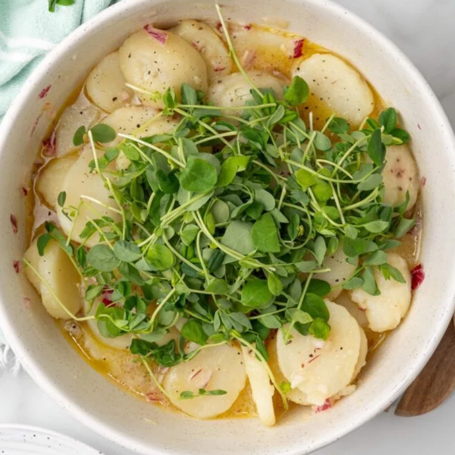 A top-down view of a white ceramic bowl filled with sliced potatoes in a light yellow broth, topped with a generous mound of fresh green microgreens.