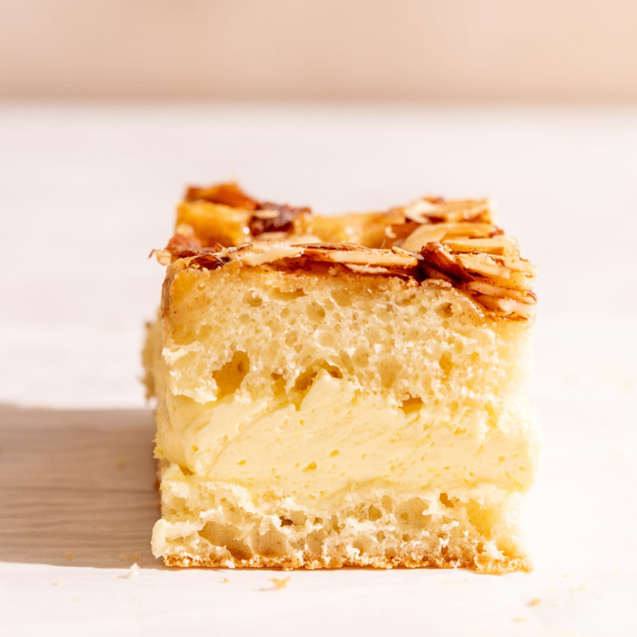 A square slice of a layered dessert, likely a cake or pastry, with a pale cream filling, a golden-brown crumb base, and toasted almond flakes on top, set against a warm beige background.