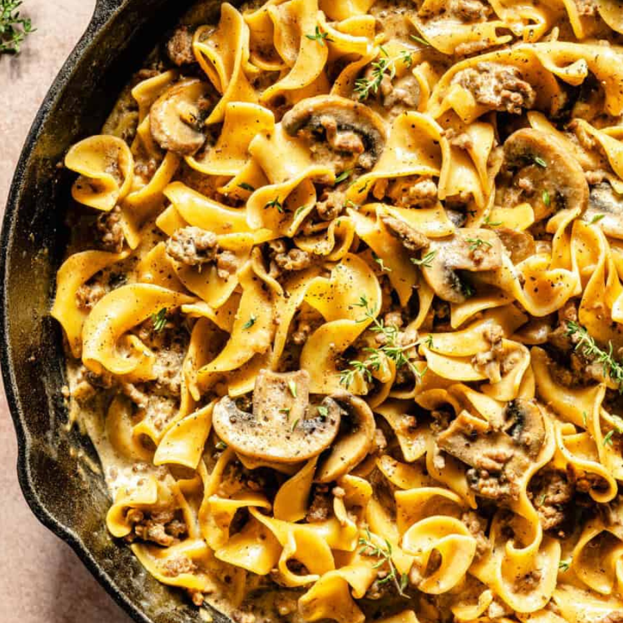 Overhead close-up of creamy mushroom pasta in a black cast iron skillet, with eggplant noodles, sliced mushrooms, browned sausage, and small thyme sprigs.