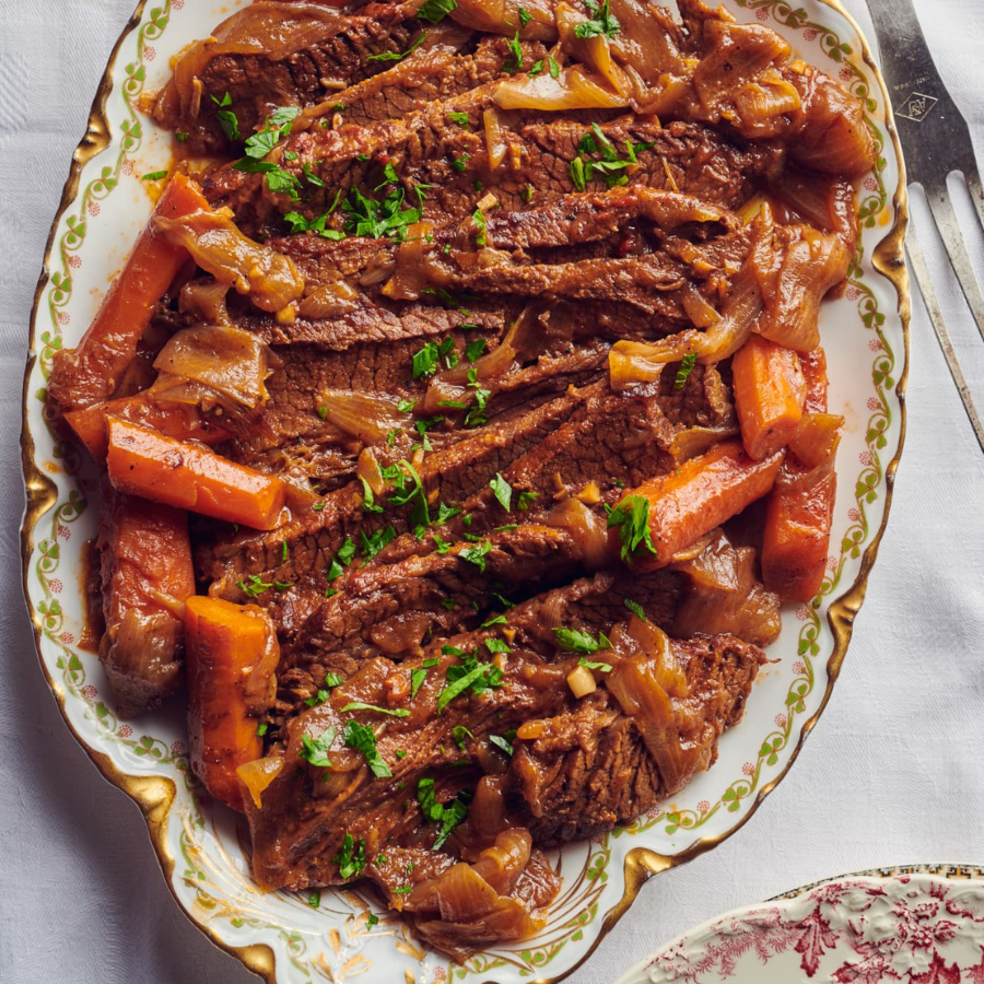 A close-up overhead shot of sliced beef stew with carrots and onions in a rich brown gravy, garnished with chopped green herbs, served on an oval white ceramic platter with a gold patterned rim.