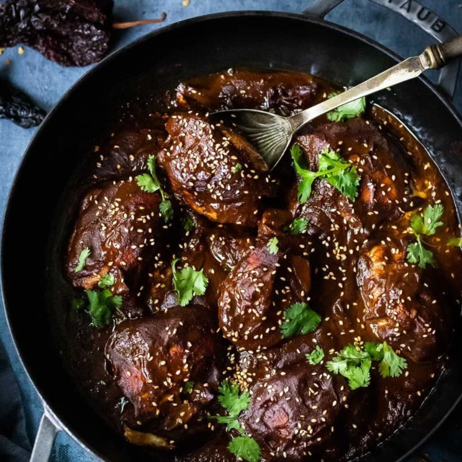 Overhead close-up of a dark, glossy braised meat dish in a black skillet, garnished with sesame seeds and fresh cilantro, with a metal serving spoon resting in the food.