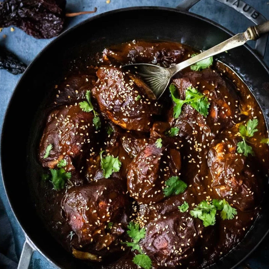 Overhead close-up of a dark, glossy braised meat dish in a black skillet, garnished with sesame seeds and fresh cilantro, with a metal serving spoon resting in the food.