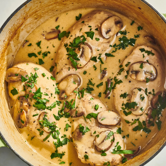 Overhead close-up of creamy mushroom chicken in a light brown sauce, topped with chopped fresh green parsley, inside a dark enameled cast iron pot.