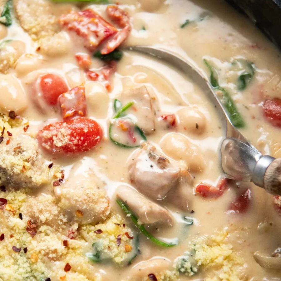 A close-up overhead shot of a creamy chicken and mushroom soup with white beans, red tomatoes, green onions, and a spoon resting in the broth.