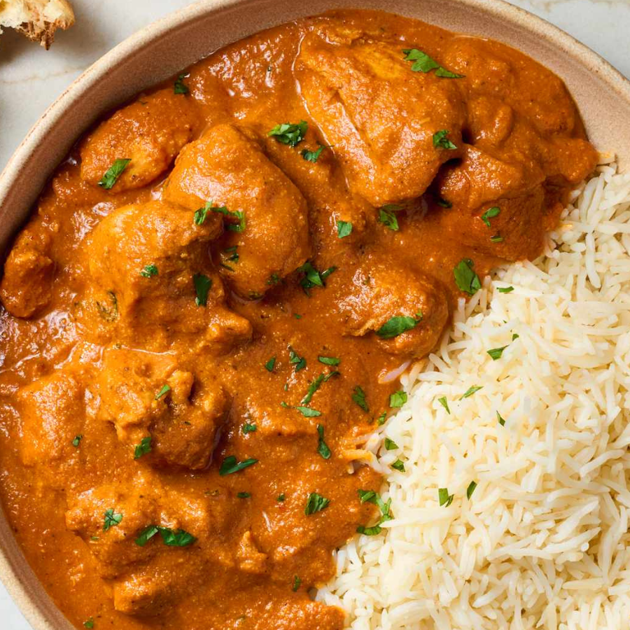 A close-up overhead shot of a bowl of chicken curry with a rich orange-red sauce, served over white rice and topped with chopped green herbs.