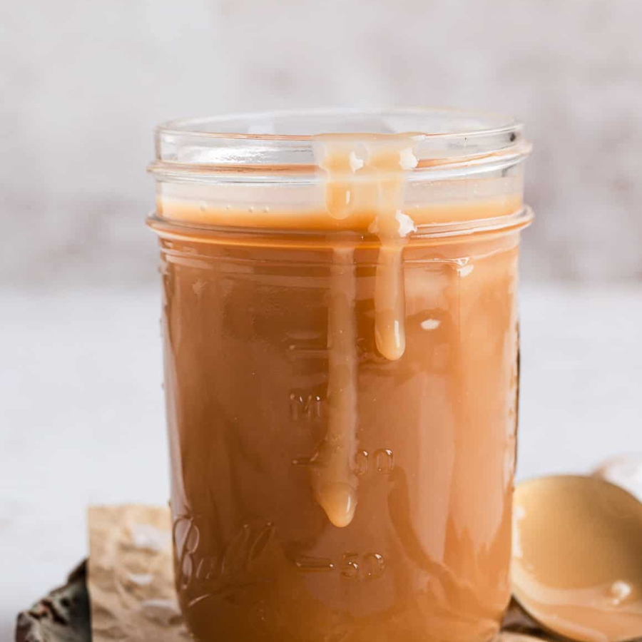 A clear glass mason jar filled with smooth caramel sauce, with caramel dripping down the front. A small spoon with caramel sits on the side, and the jar is photographed against a soft light gray background.