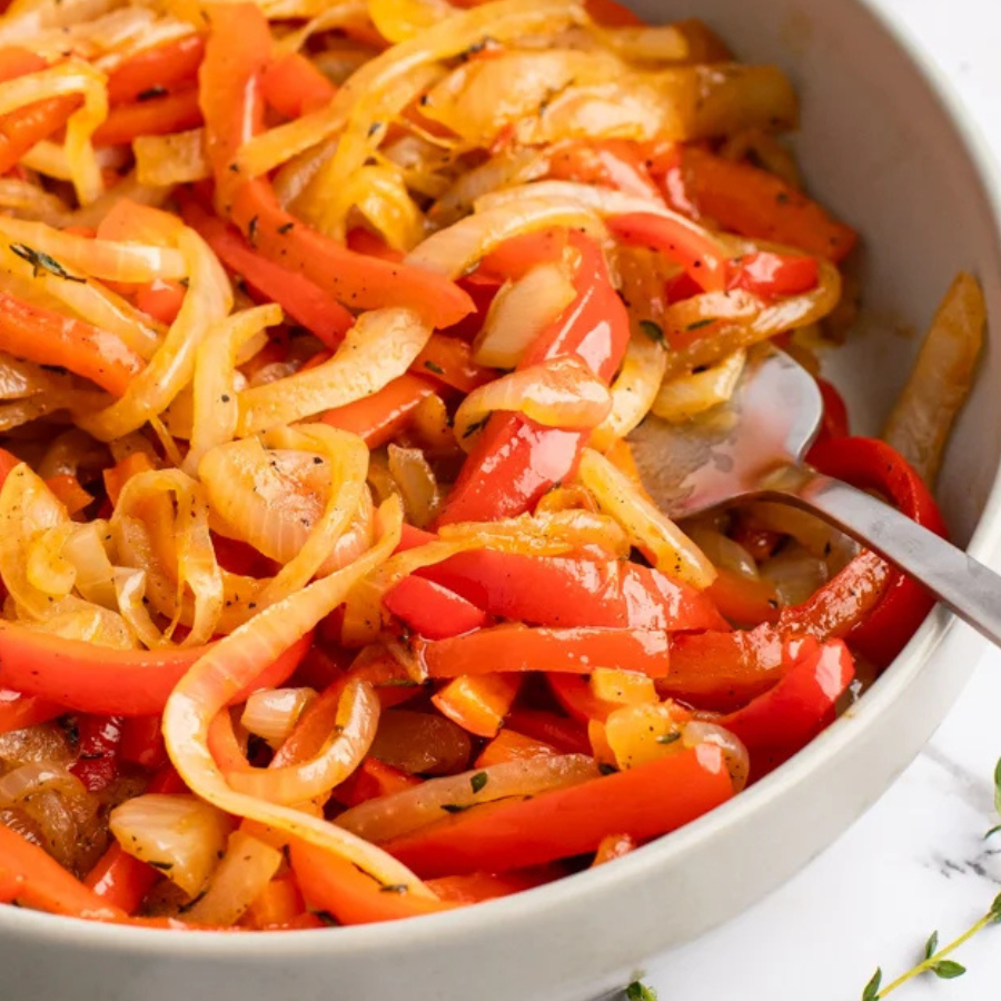 A close-up of a white bowl filled with sautéed red bell peppers and onions, with a metal spoon resting in the food.