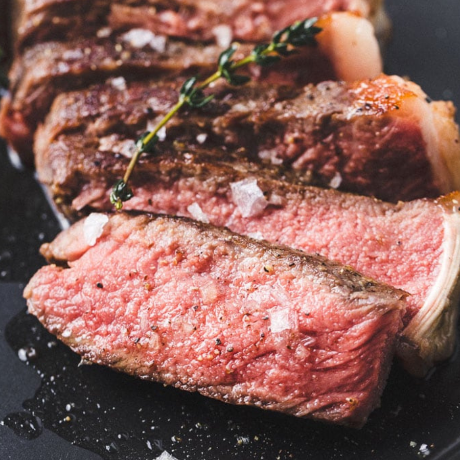 Close-up of sliced cooked beef with a pink center, browned exterior, coarse salt crystals, and a sprig of herb on a dark plate.