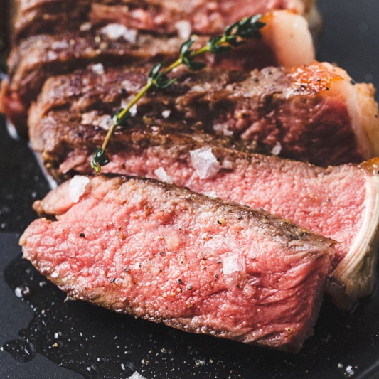 Close-up of sliced cooked beef with a pink center, browned exterior, coarse salt crystals, and a sprig of herb on a dark plate.