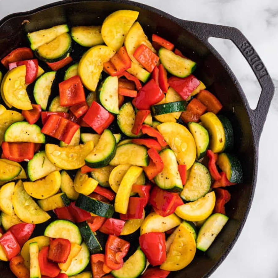 Overhead view of a cast iron skillet filled with roasted summer vegetables including yellow zucchini, red bell pepper, and green zucchini on a white marble surface.