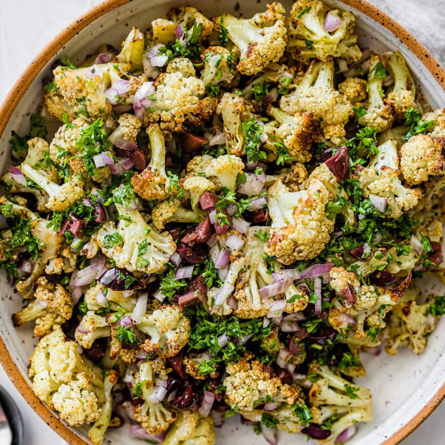A close-up overhead shot of a roasted cauliflower salad in a speckled ceramic bowl, mixed with chopped red onion, parsley, and olives.