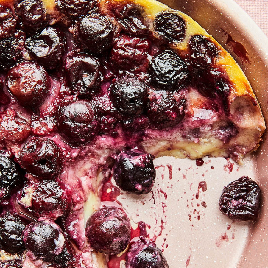 A close-up of a baked berry pie or tart with dark purple-blue cherries on top and a golden crust edge, with one slice cut out and a few cherries resting in the pinkish-brown pie dish.