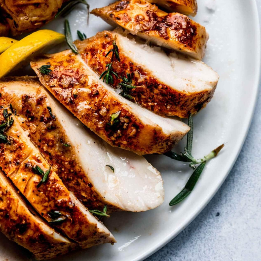 Sliced grilled chicken breast with a golden-brown crispy skin, garnished with fresh rosemary, served on a white plate with a lemon wedge in the background.