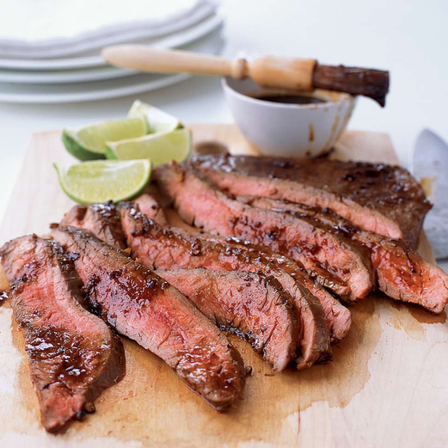 Sliced grilled steak arranged on a wooden cutting board with lime wedges and a small bowl of sauce in the background.