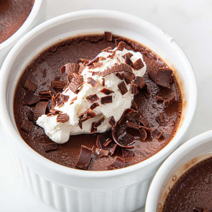 A close-up of a chocolate pudding or crème brûlée served in a white ramekin, topped with a swirl of whipped cream and chocolate shavings, on a light neutral background.