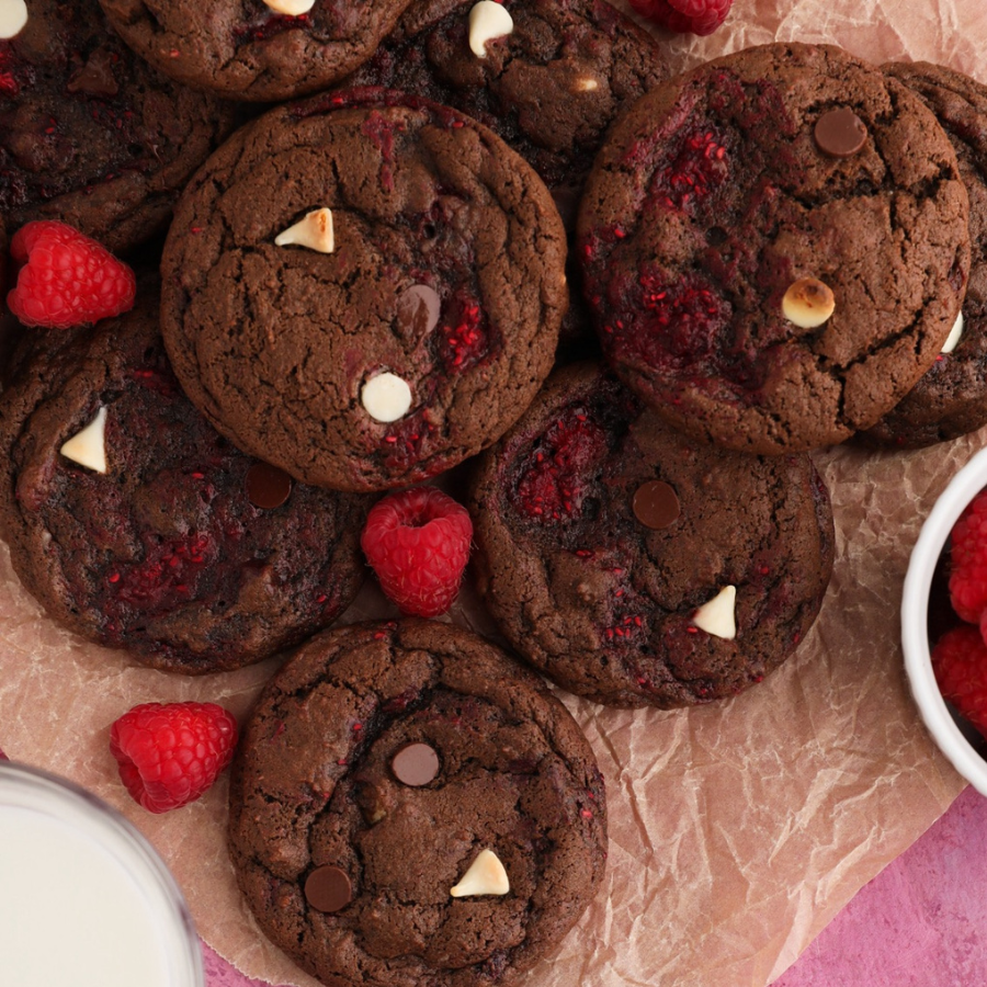 A close-up overhead shot of several chocolate chip cookies mixed with raspberries, white chocolate chips, and dark chocolate chips, arranged on brown paper with a small bowl of raspberries and a glass of milk partially visible at the edges.