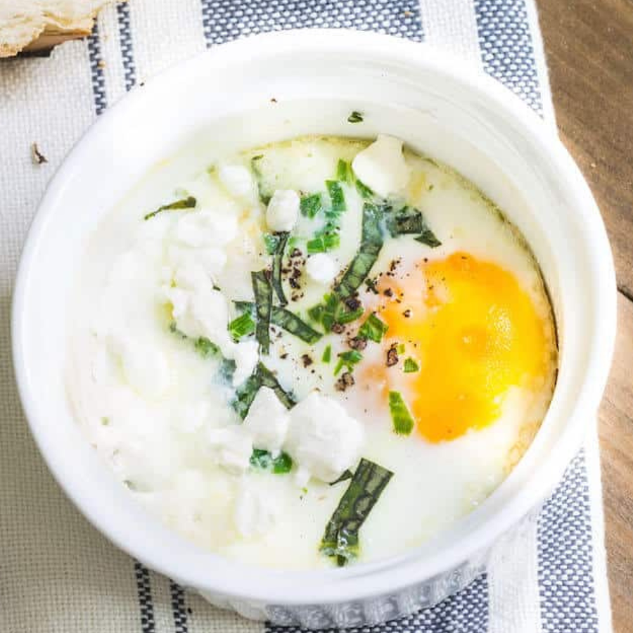 A top-down view of a cooked egg with a bright yellow yolk, white curds, chopped green herbs, and black pepper served inside a small white ceramic ramekin on a striped cloth.
