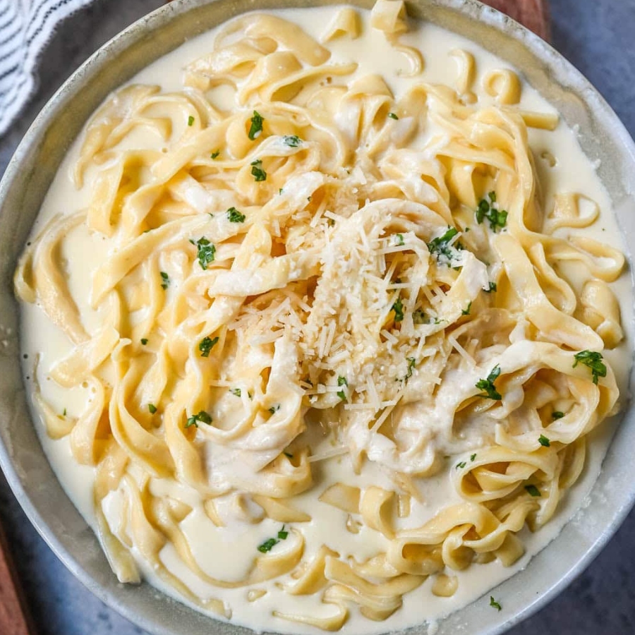 Creamy fettuccine pasta in a light gray bowl, topped with shredded cheese and chopped green herbs, photographed from above.