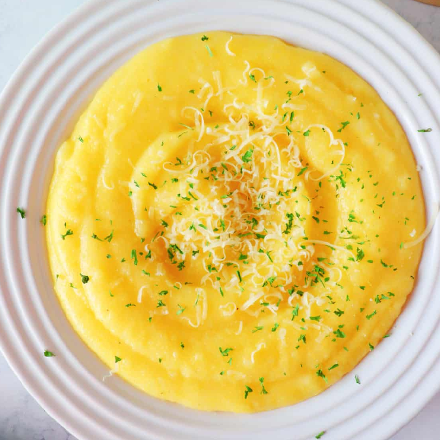 A top-down view of a creamy yellow mashed potato or polenta dish on a white plate, garnished with shredded cheese and chopped green herbs.