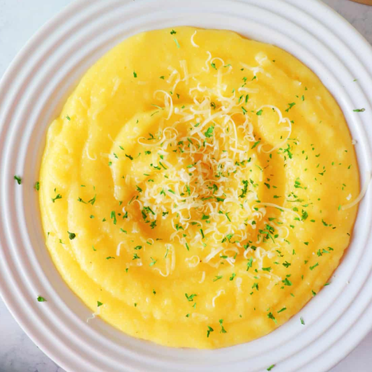 A top-down view of a creamy yellow mashed potato or polenta dish on a white plate, garnished with shredded cheese and chopped green herbs.
