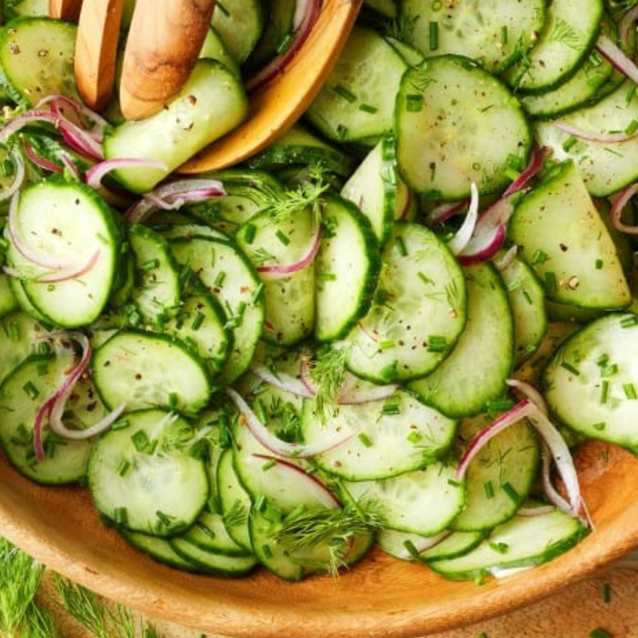 A close-up of a fresh cucumber salad in a wooden bowl, featuring thinly sliced cucumbers, chopped dill, and red onion, with wooden salad servers partially visible at the top.