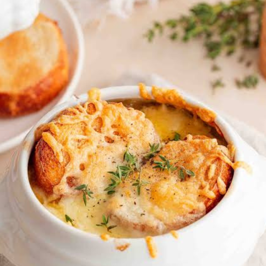 A close-up of a white ceramic bowl filled with creamy onion soup topped with melted cheese and fresh thyme, with a slice of toasted bread on a plate in the background.