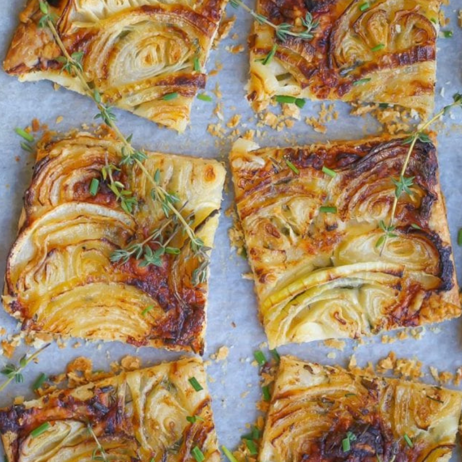 Sliced baked pastry with layered onion or pear slices, topped with herbs and crumb crumbs, arranged on a baking sheet.