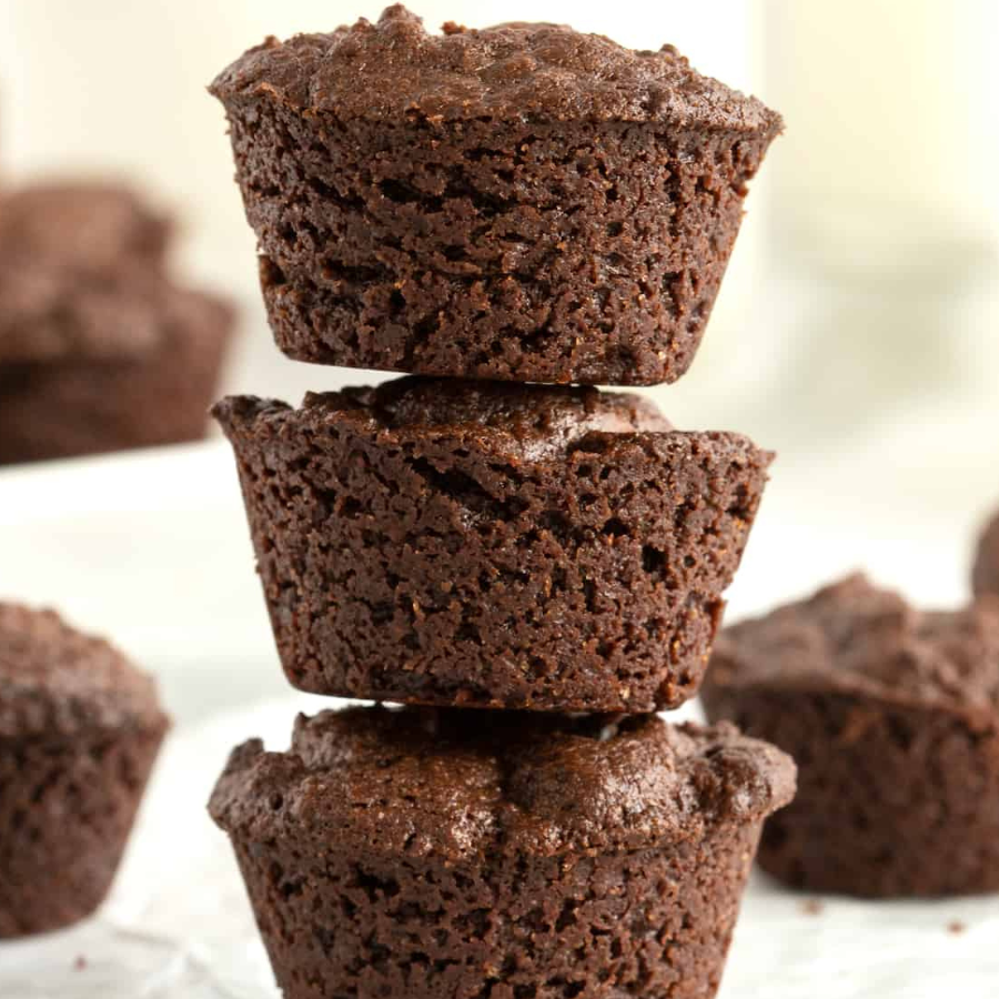 Three stacked chocolate chip muffins on a white surface with blurred chocolate muffins in the background.