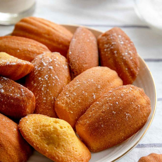 A close-up of several golden-brown madeleines on a white plate, lightly dusted with powdered sugar, set on a light table surface.