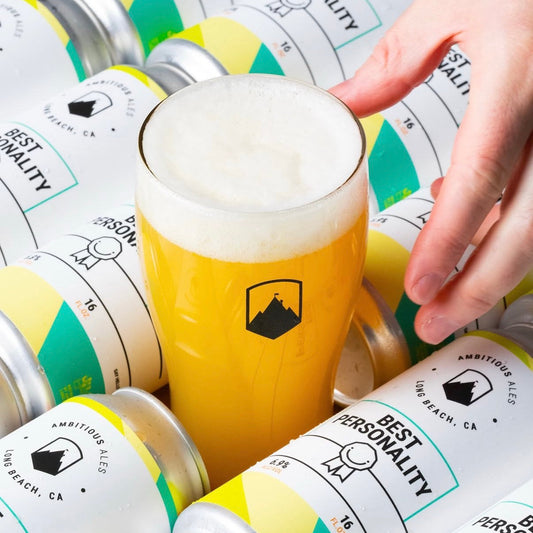 A hand holds a frothy glass of amber beer centered among many cans of Ambitious Ale 'Best Personality' beer. The cans have white labels with green and yellow geometric accents and black text.
