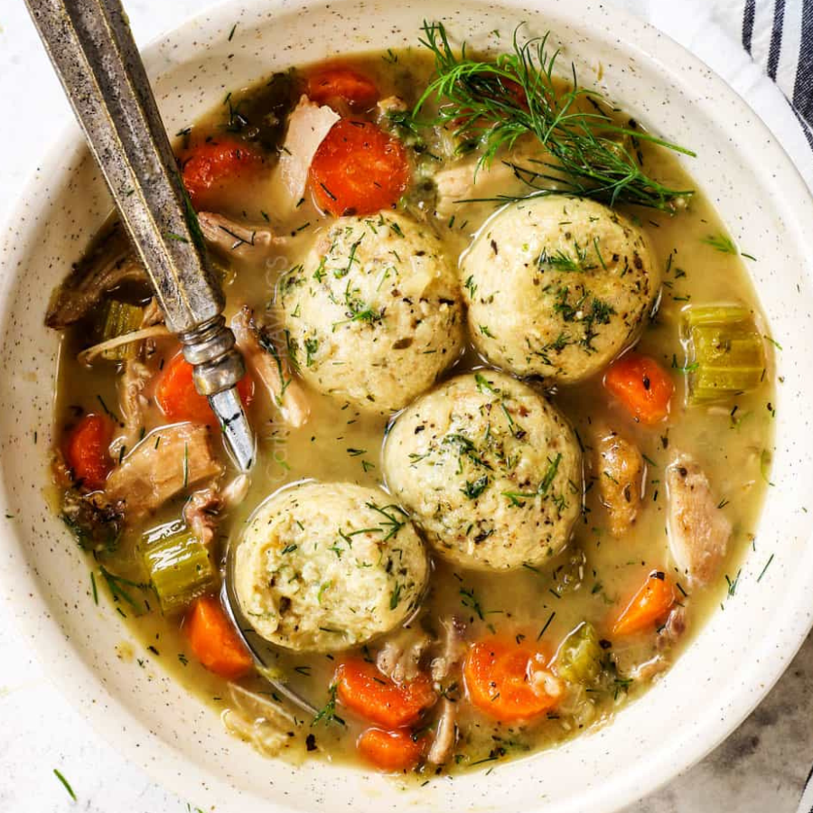 A top-down view of a bowl of chicken and dumpling soup with four large dumplings, carrots, celery, and dill in a light broth, served in a speckled cream-colored bowl with a metal spoon.