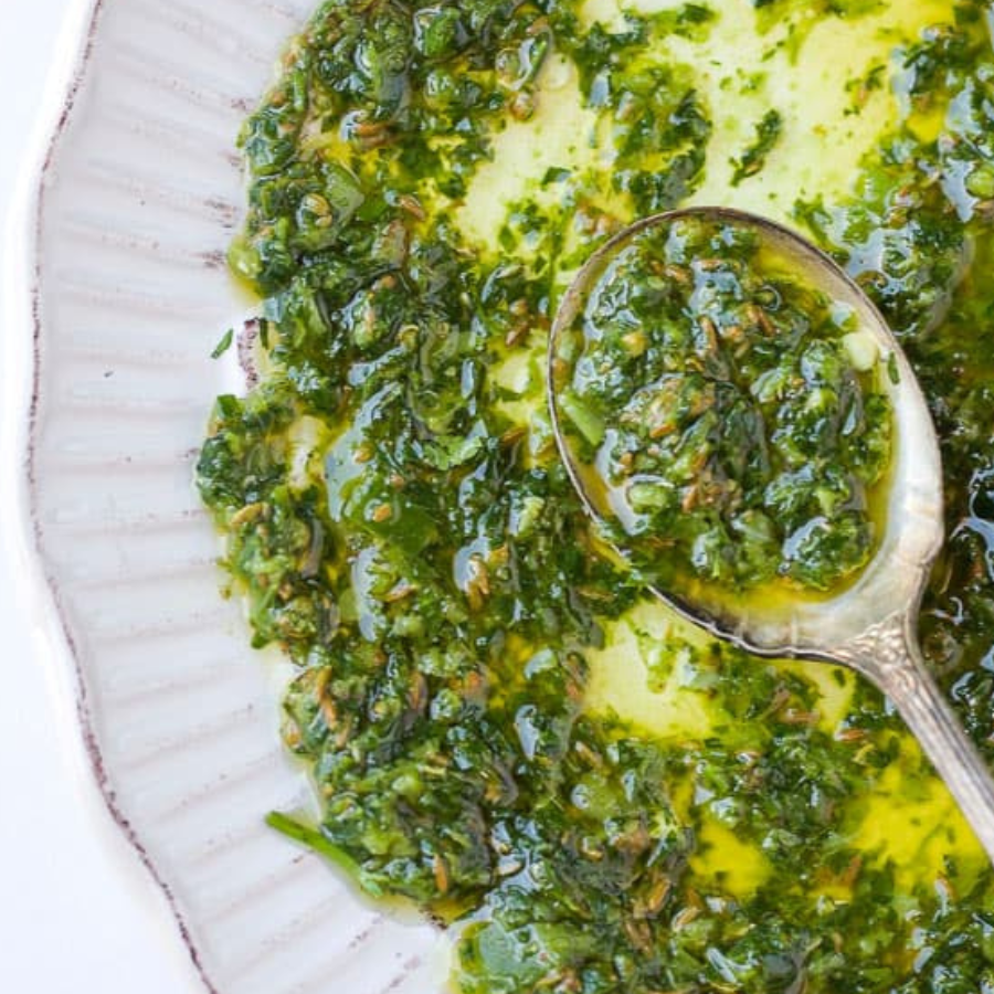 A close-up overhead shot of a green herb oil sauce or dressing in a white plate, with a metal spoon holding some of the mixture.