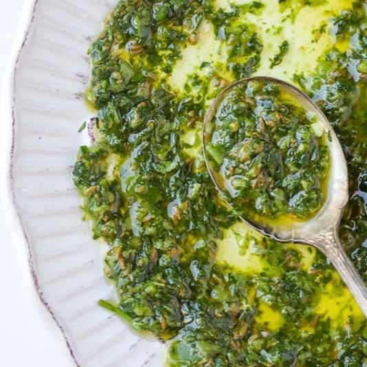 A close-up overhead shot of a green herb oil sauce or dressing in a white plate, with a metal spoon holding some of the mixture.