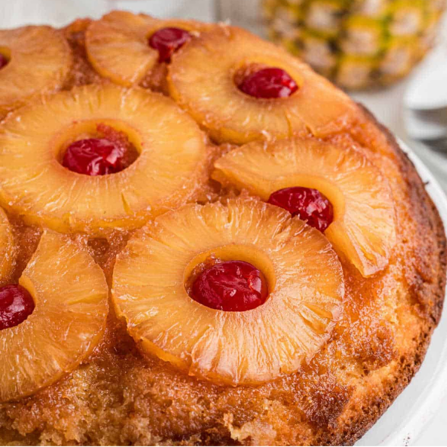 A close-up of a pineapple upside-down cake topped with glossy pineapple rings and red cherries, with a blurred pineapple in the background.