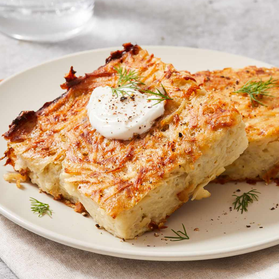 Two slices of baked potato casserole with melted cheese on a white plate, garnished with a dollop of sour cream, black pepper, and dill.