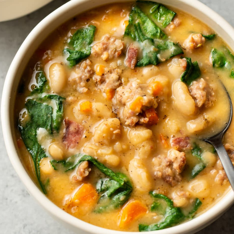 A close-up overhead shot of a white bowl filled with a hearty soup containing white beans, green spinach leaves, orange carrots, and brown ground meat in a creamy yellow broth, with a metal spoon resting on the right side.