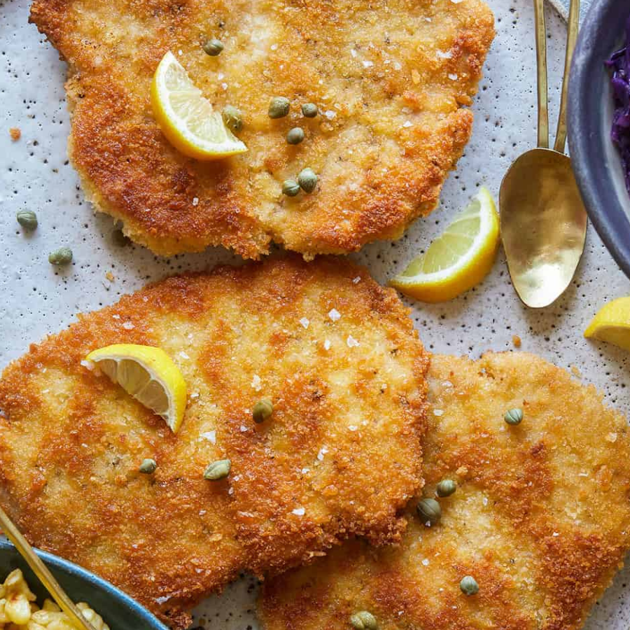 Three golden-brown breaded cutlets topped with capers and lemon wedges on a speckled light gray plate, with gold spoons and partial bowls visible around the edges.
