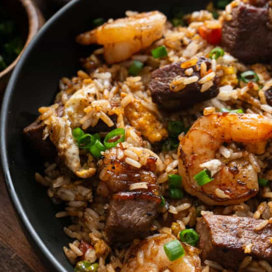 A close-up of a black bowl filled with shrimp and beef fried rice, garnished with chopped green onions, set on a wooden surface.