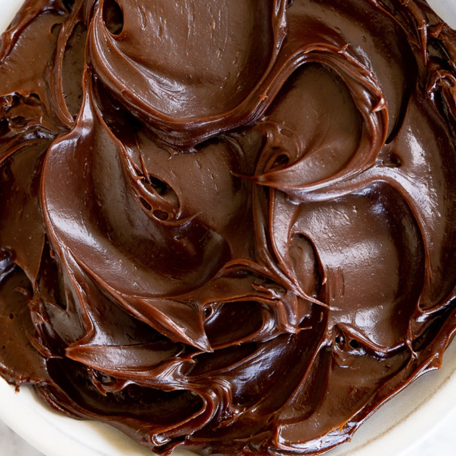 A close-up of rich, glossy chocolate frosting or whipped cream swirled in a white bowl.